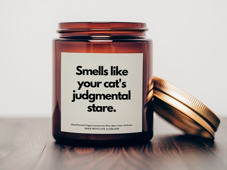 May include: A brown glass candle jar with a gold lid. The label reads "Smells like your cat's judgmental stare." Hand Poured | Vegan Coconut Soy Wax | Burn Time: 50 Hours MADE WITH LOVE & SARCASM