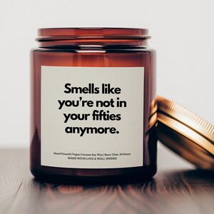 May include: A brown glass candle jar with a white label that reads "Smells like you're not in your fifties anymore." The candle is hand-poured with vegan coconut soy wax and has a burn time of 50 hours. The jar has a gold lid.