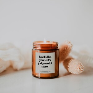 May include: A brown glass candle jar with a white label that reads "Smells like your cat's judgmental stare." The candle is lit and the flame is visible. The candle is sitting on a white surface with dried flowers.
