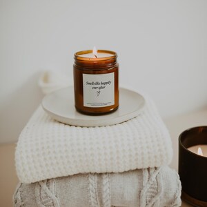May include: A brown glass candle with a white label that reads "Smells like happily ever after" with a heart symbol. The candle is sitting on a white plate on top of a stack of white knitted blankets.
