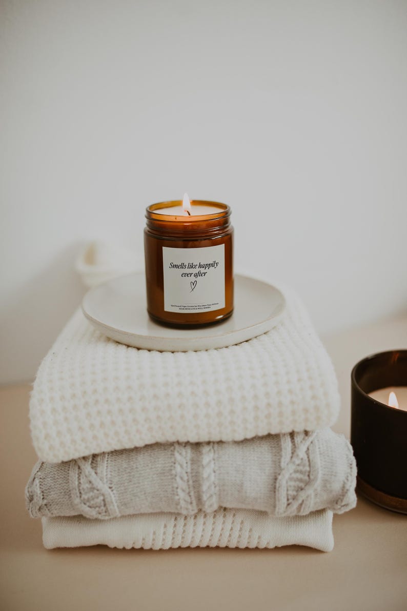 May include: A brown glass candle with a white label that reads "Smells like happily ever after" with a heart symbol. The candle is sitting on a white plate on top of a stack of white knitted blankets.