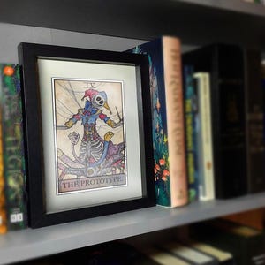 Puede incluir: Ilustración enmarcada de una figura tipo bufón esquelético sosteniendo un cuchillo. La obra de arte está etiquetada como "THE PROTOTYPE" y se exhibe en una estantería con libros.