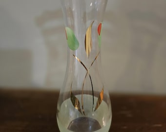 May include: A clear glass vase with a flared top and a rounded base. The vase is decorated with gold lines and green and red floral accents. The bottom of the vase has a frosted appearance.