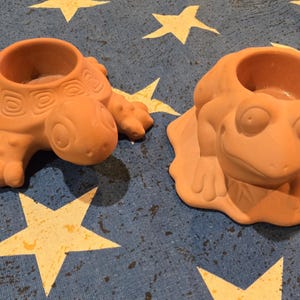 May include: Two terracotta candle holders, one shaped like a turtle and the other like a frog. The turtle has a detailed shell design, and the frog has large eyes. Both have a circular opening for a candle.