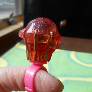 May include: A pink plastic ring featuring a large, red, gem-shaped top. The gem has gold-coloured accents and sparkles. The ring is on a finger.
