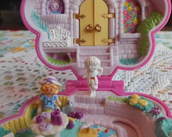 Può includere: Una casetta di giocattolo in plastica rosa con un tetto bianco e una porta gialla. La casa ha un piccolo giardino con una recinzione bianca e un piccolo stagno. Ci sono due piccole bambole all'interno della casa.