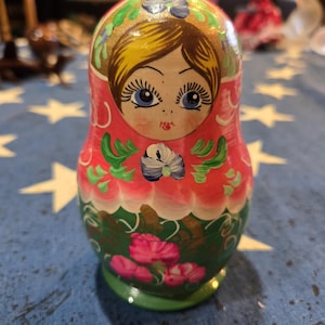 May include: A hand-painted Russian nesting doll, featuring a woman's face with blue eyes, blonde hair, and a pink blush. The doll's body is decorated with pink, green, and gold floral patterns. The base is green.