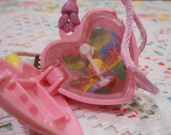 May include: A pink heart-shaped plastic locket pendant on a matching pink cord. The locket is open, revealing a clear plastic window displaying a colorful illustration of a figure on a swing set against a background of trees and butterflies.