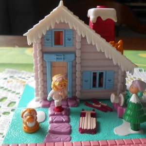Puede incluir: Un conjunto de juego de pl&aacute;stico en miniatura que presenta una caba&ntilde;a de invierno con un techo nevado, una peque&ntilde;a figura con cabello rubio, un perro, un &aacute;rbol de Navidad y un trineo. La caba&ntilde;a tiene una puerta y ventanas azules.