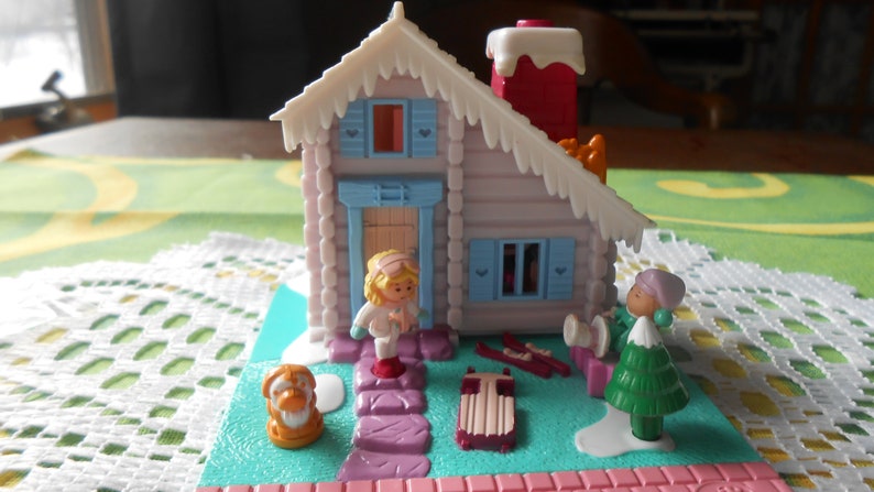 Puede incluir: Un conjunto de juego de pl&aacute;stico en miniatura que presenta una caba&ntilde;a de invierno con un techo nevado, una peque&ntilde;a figura con cabello rubio, un perro, un &aacute;rbol de Navidad y un trineo. La caba&ntilde;a tiene una puerta y ventanas azules.