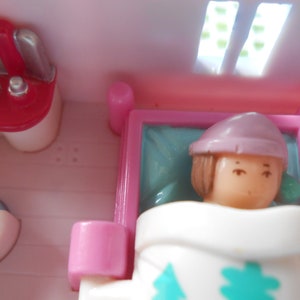 Puede incluir: Un juego de casa de mu&ntilde;ecas rosa y blanco con una mu&ntilde;eca con un sombrero rosa y una manta blanca acostada en una cama. La casa de mu&ntilde;ecas tambi&eacute;n cuenta con un tocador con un espejo y una silla peque&ntilde;a.