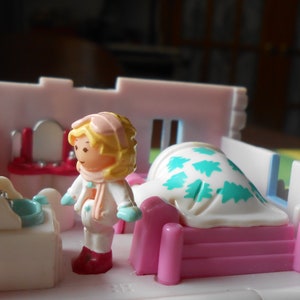 Puede incluir: Un juego de casa de mu&ntilde;ecas de pl&aacute;stico blanco con una cama rosa y blanca, un tocador blanco con espejo y una mu&ntilde;eca que lleva un atuendo de invierno blanco con un gorro rosa y guantes azules.