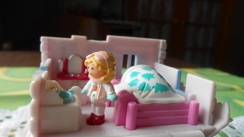 Puede incluir: Un juego de casa de mu&ntilde;ecas de pl&aacute;stico blanco con una cama rosa y blanca, un tocador blanco con espejo y una mu&ntilde;eca que lleva un atuendo de invierno blanco con un gorro rosa y guantes azules.