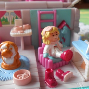 Puede incluir: Un juego de casa de mu&ntilde;ecas de pl&aacute;stico rosa y blanco con una mu&ntilde;eca, un perro y muebles. La casa de mu&ntilde;ecas tiene una chimenea azul y blanca, una cama rosa y blanca y una mesa con una silla. La mu&ntilde;eca lleva un abrigo blanco y un gorro rojo. El perro es marr&oacute;n y blanco.