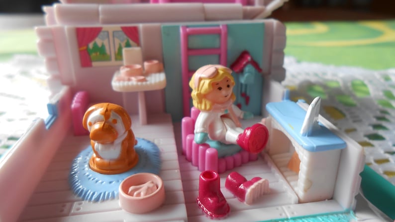 Puede incluir: Un juego de casa de mu&ntilde;ecas de pl&aacute;stico rosa y blanco con una mu&ntilde;eca, un perro y muebles. La casa de mu&ntilde;ecas tiene una chimenea azul y blanca, una cama rosa y blanca y una mesa con una silla. La mu&ntilde;eca lleva un abrigo blanco y un gorro rojo. El perro es marr&oacute;n y blanco.