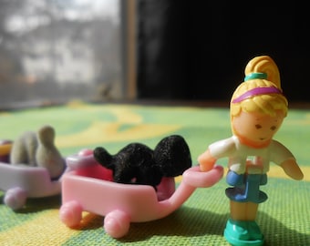 Può includere: Una bambola di una bambina giocattolo con i capelli biondi e una fascia viola tira un carrello rosa con un cane giocattolo nero dentro. Il carrello ha le ruote e una maniglia. La bambola indossa una camicia bianca e jeans blu.