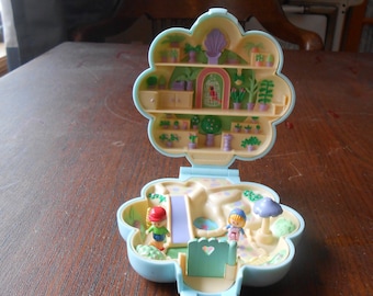 May include: A vintage, open, flower-shaped playset with a light blue exterior. The interior features miniature figures, a slide, and a garden scene with small plants. The top section has shelves with more miniature plants and a small birdcage.