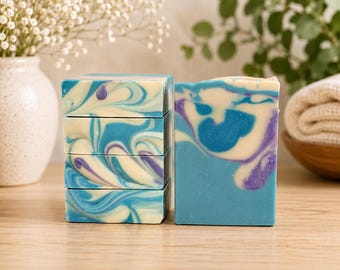 May include: A stack of four rectangular soap bars and one upright soap bar featuring a marbled pattern of cream, bright blue, and purple swirls. The soap bars have a smooth, solid texture and are arranged on a light-coloured wooden surface.