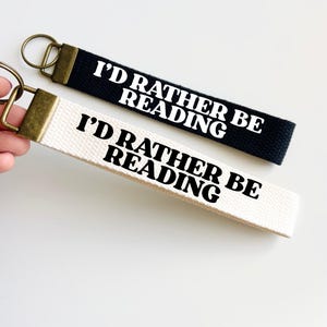Pode incluir: Dois chaveiros com o texto "I'D RATHER BE READING". Um é preto, o outro branco. Ambos têm um anel e um fecho de metal dourado.