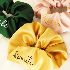 May include: Three satin scrunchies in different colors with names embroidered on them. The scrunchies are green, pink, and yellow. The names are "Akvile", "Rima", and "Rimute".