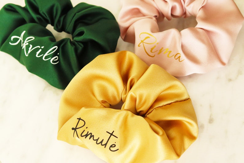 May include: Three satin scrunchies in different colors with names embroidered on them. The scrunchies are green, pink, and yellow. The names are "Akvile", "Rima", and "Rimute".