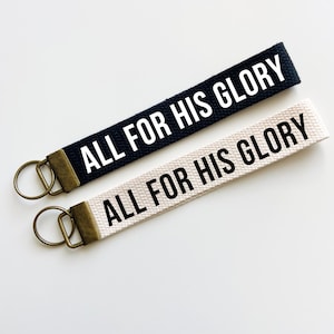 Könnte beinhalten: Zwei Schlüsselanhänger mit dem Text "ALL FOR HIS GLORY" darauf gedruckt. Ein Schlüsselanhänger ist schwarz mit weißem Text und der andere ist weiß mit schwarzem Text.