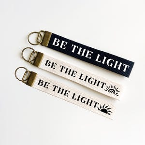 May include: Three keychains with the text "Be the Light" and a sun graphic. The top keychain is black with white text, the middle keychain is white with black text, and the bottom keychain is white with black text and a sun graphic.