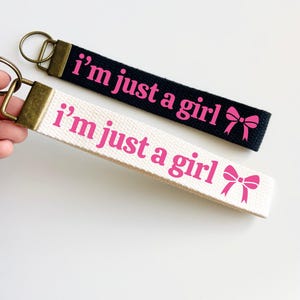 May include: Two keychains with the text "i'm just a girl" in pink lettering. One keychain is black with a pink bow and the other is white with a pink bow.