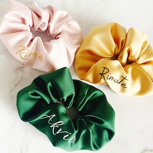 May include: Three satin scrunchies in different colors: pink, yellow, and green. Each scrunchie has a name written on it in gold or white lettering: "Rima", "Rimute", and "Akvii".