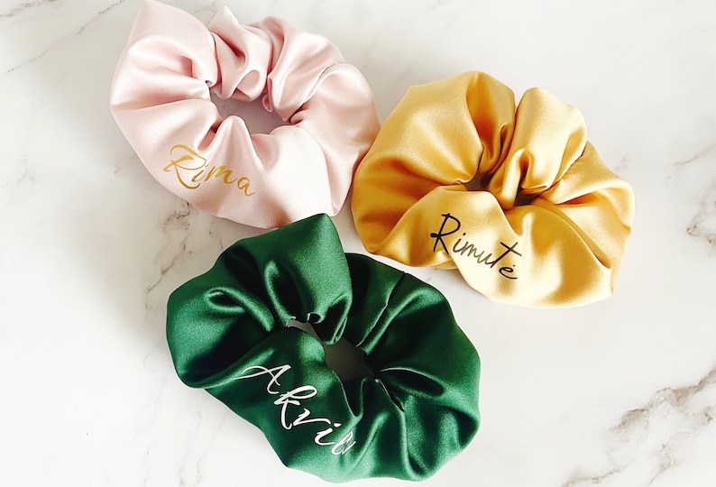 May include: Three satin scrunchies in different colors: pink, yellow, and green. Each scrunchie has a name written on it in gold or white lettering: "Rima", "Rimute", and "Akvii".