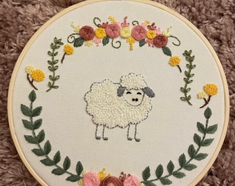 May include: Embroidered hoop art featuring a white sheep with a gray face and black eyes, surrounded by a wreath of green leaves and pink, yellow, and orange flowers.