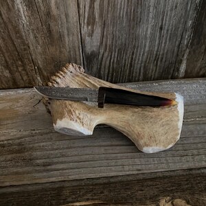 May include: A handcrafted knife with a patterned blade and a dark, curved handle. The knife rests on a piece of antler, set against a rustic wooden background. The blade appears to be approximately 6 inches long.