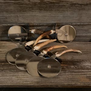 May include: A set of six pizza cutters with antler handles. The cutters have a silver finish and are arranged in a pile on a wooden surface.