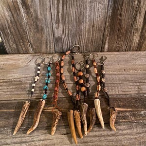 May include: A collection of keychains featuring antler pendants. The keychains have various beaded accents in colors like turquoise, brown, and white. Each keychain has a metal ring for attaching keys.