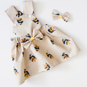 May include: A beige linen dress with a yellow and black bumblebee print. The dress has a matching yellow bow and adjustable straps. It comes with a separate matching yellow bow headband.