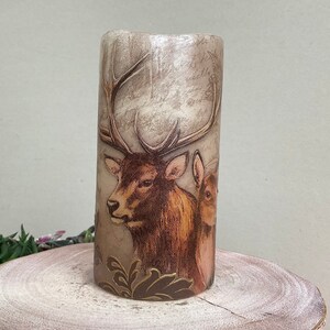 May include: A beige pillar candle with a brown and white image of a deer and a rabbit. The candle has a gold and brown floral design at the bottom.