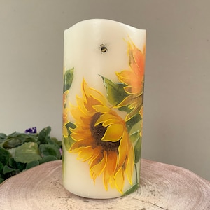 以下が含まれることがあります： ひまわりとミツバチが描かれた白い円柱形のキャンドル。ひまわりは黄色で、中心は茶色、葉は緑色です。