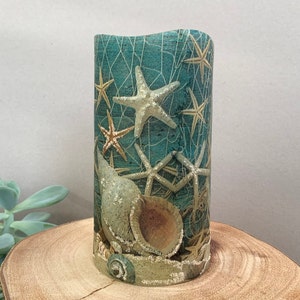 May include: A teal blue pillar candle with a seashell and starfish design. The candle has a textured surface and is decorated with gold glitter.
