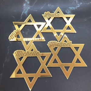 May include: Four gold-colored Star of David decorations with the names Mia, Laura, Michael, and Michelle.