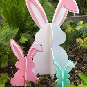 May include: Three wooden bunny cutouts in different sizes. The largest bunny is white with pink ears, the middle bunny is pink with white ears, and the smallest bunny is light green with white ears. The bunnies are standing in a garden setting.
