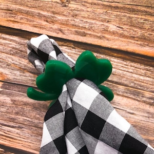 May include: Green shamrock-shaped napkin ring made of plastic. The ring is holding a black and white checkered napkin.