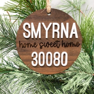 May include: A round wooden ornament with the text "SMYRNA home sweet home 30080" in white lettering. The ornament is hanging from a brown twine string.