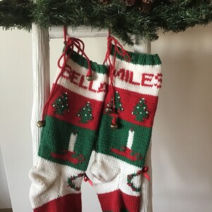 May include: Two red, white, and green Christmas stockings with the words "BELLE" and "MILES" knitted into the fabric. The stockings have a green, white, and red striped pattern with a small Christmas tree and candle design. The stockings are hanging on a white ladder.