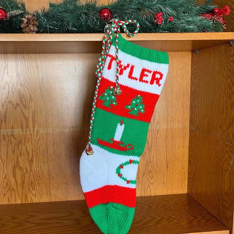 May include: A green, red, and white knitted Christmas stocking with the name "Tyler" embroidered on the front. The stocking has a green and white rope with bells attached.
