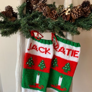 May include: Two red, white, and green knitted Christmas stockings with the names "Jack" and "Katie" embroidered on them. The stockings are decorated with small green Christmas trees and white candles.