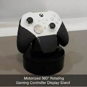 Puede incluir: Un mando de videojuegos blanco y negro sobre un soporte de exhibición giratorio motorizado de 360 grados de color negro. El mando tiene joysticks y botones negros. El soporte tiene el texto "Motorized 360° Rotating Gaming Controller Display Stand".