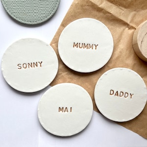 May include: Four white ceramic coasters with gold lettering. The coasters say "Mummy", "Sonny", "Daddy", and "Mai".