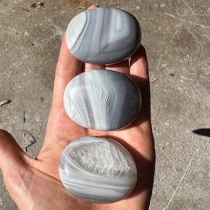 Pode incluir: Três pedras de ágata lisas de forma oval com bandas brancas e cinzas. As pedras são polidas e têm um aspeto natural e orgânico.