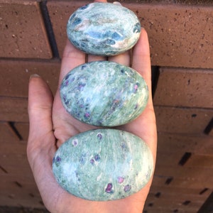 May include: Three oval stones with a green and blue base colour, speckled with small red and pink inclusions. The stones are smooth and polished, and are held in a hand. The stones are likely used for decorative or healing purposes.