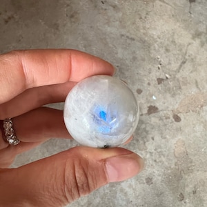以下が含まれることがあります： 青い虹色の光沢を持つ白いムーンストーンの球体。球体は滑らかで磨かれています